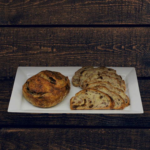 A plate with sliced and whole round loaves of fig & roasted walnut bread boule- one is a round with visible nuts and figs, and the other is a sliced loaf with similar inclusions, against a wooden background.