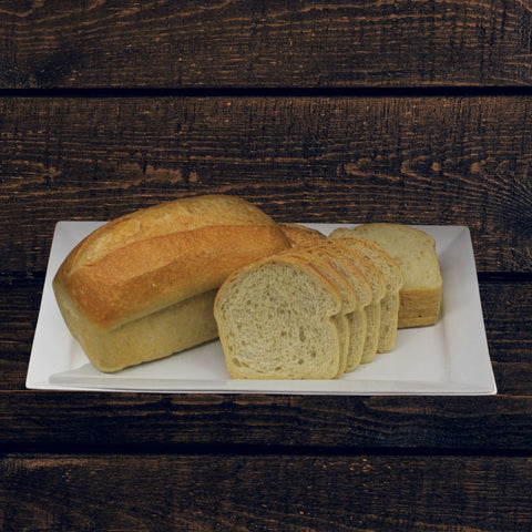A loaf of artisan white sandwich bread called Il Forno on a white plate, with sliced bread in the background, placed against a wooden door backdrop.