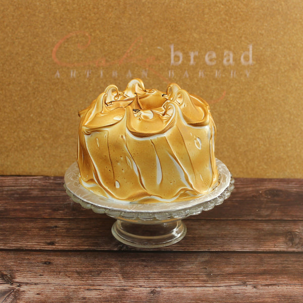 A golden-brown lemon meringue layer cake with toasted meringue on top, presented on a glass cake stand.