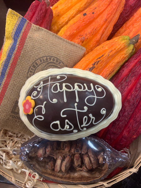 A personalized chocolate Easter egg with 'Happy Easter' written on it, surrounded by various items like dried cacao pods and a basket.