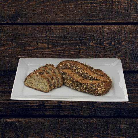 A rustic-looking batard loaf of artisan multigrain seed bread on a plate, with visible seeds and a golden-brown crust, placed on a wooden surface.
