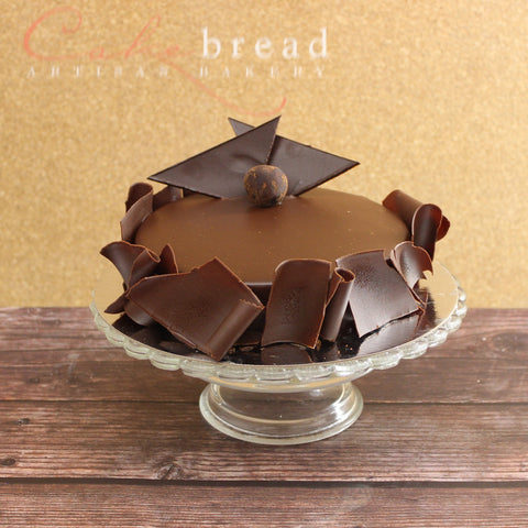 A round flourless chocolate truffle cake garnished with chocolate shards and a chocolate decoration on top, presented on a glass cake stand.