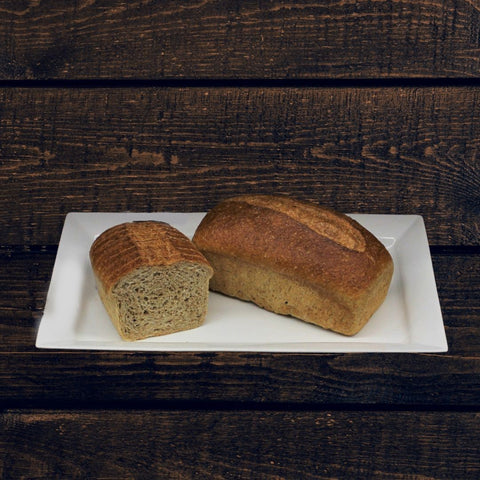 Handcrafted whole wheat sandwich bread on a white plate, with one slice cut and placed alongside the loaf. The bread has a golden-brown color, and the plate is placed against a wooden background.